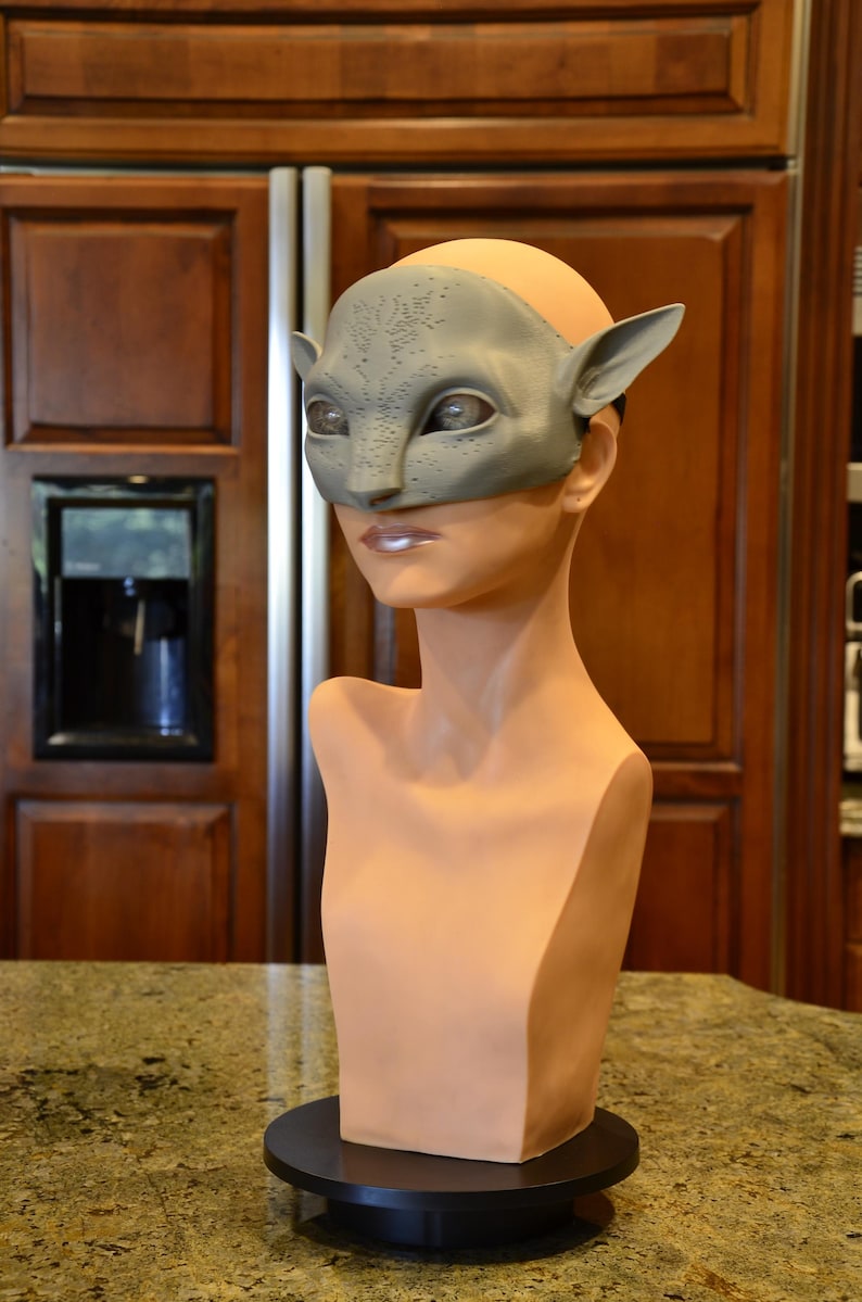 Puede incluir: Una m&aacute;scara gris texturizada inspirada en felinos con orejas puntiagudas y aberturas para los ojos, exhibida en una cabeza de maniqu&iacute; lisa y clara. La m&aacute;scara presenta un patr&oacute;n de puntos en la frente y el puente de la nariz.