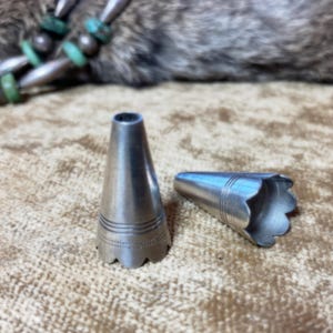 Könnte beinhalten: Zwei silberfarbene Metallkegel mit gezackten Kanten. Einer steht aufrecht, der andere liegt auf der Seite. Beide haben geätzte Details und eine konische Form. Der Hintergrund ist eine strukturierte, braune Oberfläche.