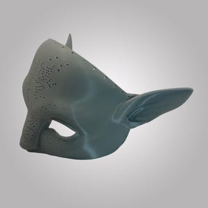 Puede incluir: Una m&aacute;scara gris de superficie lisa con orejas puntiagudas y una serie de peque&ntilde;os orificios perforados dispuestos en un patr&oacute;n org&aacute;nico ramificado en la parte frontal. La m&aacute;scara tiene una abertura para el ojo y forma contorneada.
