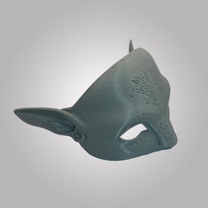 Puede incluir: Una m&aacute;scara gris mate esculpida con orejas puntiagudas y una superficie texturizada con peque&ntilde;os agujeros perforados en la frente y la nariz. La m&aacute;scara tiene una abertura para el ojo y una forma contorneada que cubre la parte superior del rostro.