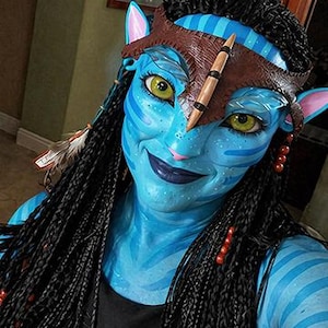 Puede incluir: Un personaje de piel azul con ojos amarillos, rayas azul oscuro y orejas puntiagudas. El personaje lleva una diadema de cuero marr&oacute;n con una punta central, cabello negro trenzado con plumas y cuentas rojas, y una prenda superior negra.