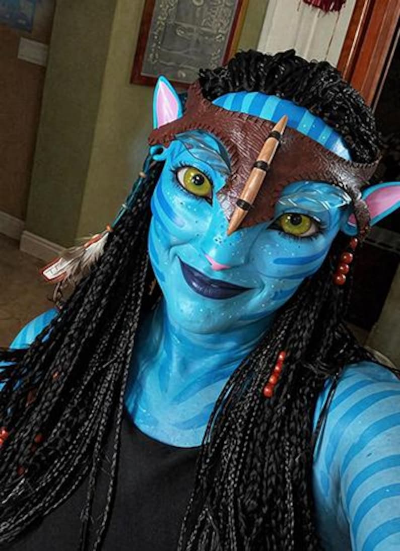 Puede incluir: Un personaje de piel azul con ojos amarillos, rayas azul oscuro y orejas puntiagudas. El personaje lleva una diadema de cuero marr&oacute;n con una punta central, cabello negro trenzado con plumas y cuentas rojas, y una prenda superior negra.
