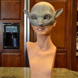 Puede incluir: Una m&aacute;scara gris texturizada inspirada en alien&iacute;genas con orejas puntiagudas y grandes aberturas para los ojos en forma de almendra. La m&aacute;scara est&aacute; colocada sobre un busto de maniqu&iacute; liso y claro que descansa sobre una base circular negra.