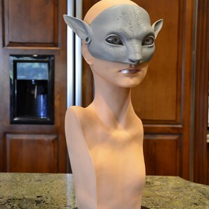 Puede incluir: Una m&aacute;scara de color gris claro, texturizada e inspirada en felinos, con orejas puntiagudas y marcas faciales detalladas, colocada sobre un busto de maniqu&iacute; de color beige con forma humana montado en una base circular negra.
