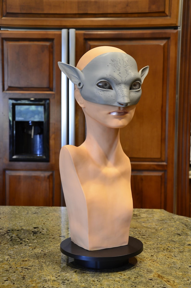 Puede incluir: Una m&aacute;scara de color gris claro, texturizada e inspirada en felinos, con orejas puntiagudas y marcas faciales detalladas, colocada sobre un busto de maniqu&iacute; de color beige con forma humana montado en una base circular negra.