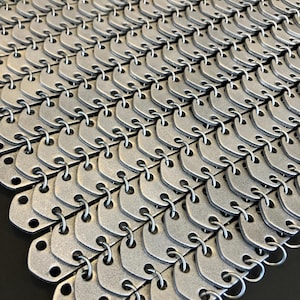 May include: A close-up of a silver chainmail, a type of armor made of interlocking metal rings. The chainmail is made of small, flat, silver-colored metal plates connected by rings.