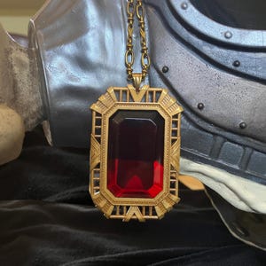 Może przedstawiać: Złoty naszyjnik z wisiorkiem z dużą, prostokątną, ciemnoczerwoną szlachetnym kamieniem. Wisiorek ma wzór Art Deco z geometrycznymi wzorami. Łańcuszek jest również w kolorze złotym. Naszyjnik jest prezentowany na ciemnym tle.