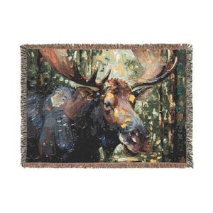 May include: A rectangular woven throw blanket featuring a detailed depiction of a moose in a forest setting. The design uses a palette of dark brown, forest green, and golden yellow tones with a visible textured weave and fringed edges on all sides.