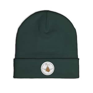 May include: A dark forest green knit beanie featuring a cuffed brim. Centered on the cuff is a circular white embroidered patch with a brown pinecone graphic and the word "Omnia" stitched in a cursive font above the pinecone illustration.