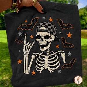 Può includere: Una t-shirt nera con la grafica di uno scheletro che indossa un berretto da baseball a scacchi. Lo scheletro fa un gesto della mano rock and roll. Il design include stelle arancioni e grigie e sagome di pipistrelli neri intorno.