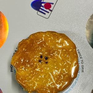May include: A close-up of a round, golden yellow phone grip with a black dot pattern in the center. The grip is made of a clear, sticky material and is designed to be attached to the back of a phone.