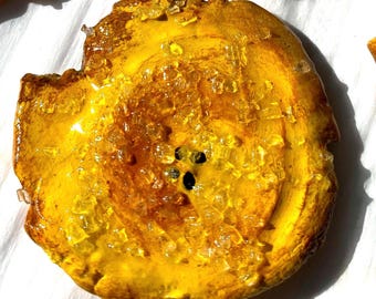 May include: Close-up of a golden-yellow, circular food item with a glossy, textured surface. The food has a dark brown center with small black spots and is sprinkled with translucent crystals. The item is on a white surface.