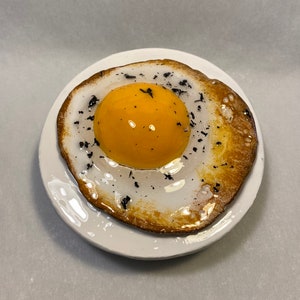 May include: A ceramic plate with a fried egg design. The egg yolk is a bright yellow, surrounded by a white egg white with a golden-brown edge. Black specks are sprinkled on the egg white.