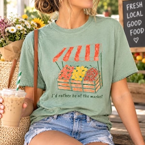Peut inclure: Un t-shirt à manches courtes vert sauge avec un motif représentant un étal de marché doté d'un auvent rayé rouge et blanc, des caisses en bois remplies de fruits rouges et jaunes, et le texte "I'd rather be at the market" en vert foncé.