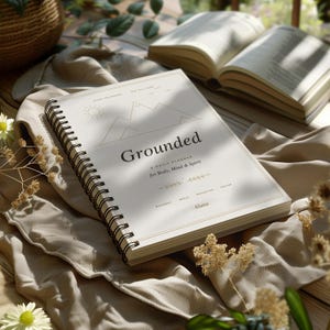 Könnte beinhalten: Ein spiralgebundener Tagesplaner mit cremefarbenem Einband, der eine Strichzeichnung von Bergen und einer Sonne zeigt. Der Text lautet Grounded, A Daily Planner for Body, Mind & Spirit sowie Aluna, mit Abschnitten für Bewegung und Mahlzeiten.