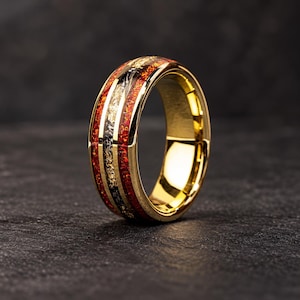 May include: A gold-toned ring featuring a central inlay composed of vibrant red crushed material and shimmering gold-coloured flecks. The band has a polished, reflective metallic finish with a smooth, rounded profile and a wide, sturdy construction.