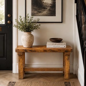 May include: A rectangular wooden console table with a natural finish, featuring a textured beige ceramic vase filled with green foliage, a dark brown wooden bowl, and two stacked books titled Live Beautifully and This Is Home.