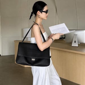 May include: A large black leather shoulder bag with a rectangular silhouette, a fold-over flap, and a silver-toned metal turn-lock closure. The bag features a single wide shoulder strap and a textured surface finish, providing a structured appearance.