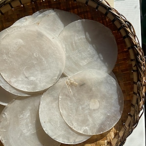 May include: A collection of translucent, circular capiz shell discs layered within a round, woven brown bamboo basket. Each thin, off-white shell piece features a natural, pearlescent sheen and a smooth, flat surface with visible organic textures.