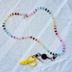 May include: A lanyard featuring a long strand of multicoloured translucent beads arranged in a gradient pattern. The lanyard includes a black retractable badge reel, a silver-toned toggle clasp, and a bright yellow plastic scissor-shaped charm.
