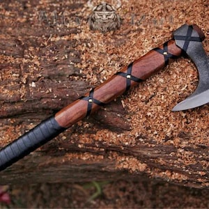 Könnte beinhalten: Eine Handaxt mit einem Kopf aus dunklem Schmiedemetall und einem Holzstiel. Der Stiel ist am unteren Ende mit schwarzem Leder umwickelt und weist ein gekreuztes Lederschnürmuster auf, das mit Metallnieten am Schaft befestigt ist.