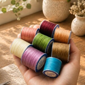 May include: A collection of nine small spools of sewing thread in various colors including cream, pink, light blue, green, brown, orange, and dark red, held together in a cluster. Each spool features a white label on the side.