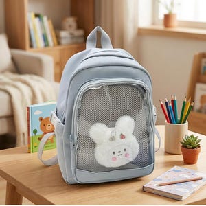 Puede incluir: Una mochila de color azul claro que cuenta con un bolsillo frontal con una ventana de malla transparente. Dentro del bolsillo de malla hay una cara de animal de peluche blanca y esponjosa con mejillas rosadas y pequeños pines decorativos.