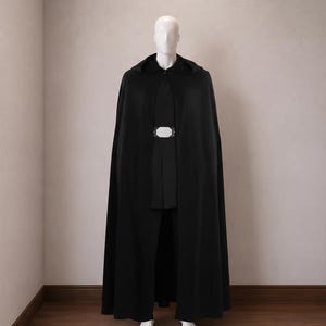 Könnte beinhalten: Ein bodenlanger schwarzer Umhang mit Kapuze über einer schwarzen Tunika und schwarzen Hose, ergänzt durch eine auffällige rechteckige silberfarbene Gürtelschnalle in der Taille. Das Set wird auf einer weißen Schaufensterpuppe präsentiert.