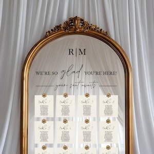 May include: An arched mirror with an ornate gold frame featuring the text R|M, WE'RE SO glad YOU'RE HERE!, and your seat awaits. Eight cream-colored table seating cards are attached to the mirror surface, each secured with a gold wax seal.