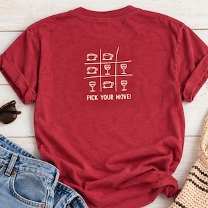 Puede incluir: Una camiseta de manga corta de color rojo oscuro con un gráfico blanco de un tablero de tres en raya. La cuadrícula contiene iconos de tazas de café y copas de vino, con el texto PICK YOUR MOVE! impreso en letras blancas.