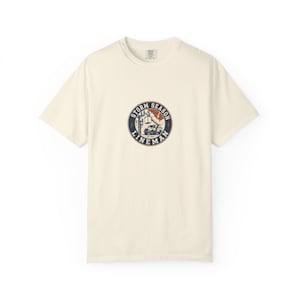 May include: A cream-coloured short-sleeve t-shirt featuring a circular graphic on the chest. The graphic depicts a utility truck with a bucket lift near a power pole, surrounded by the text "STORM SEASON LINEMAN" in a dark blue circular border.