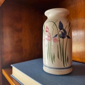 May include: A cylindrical ceramic vase with a cream-colored glaze, featuring hand-painted pink and blue floral motifs with green stems. The base is accented with a thin blue horizontal line, and the rim is slightly flared and rounded.
