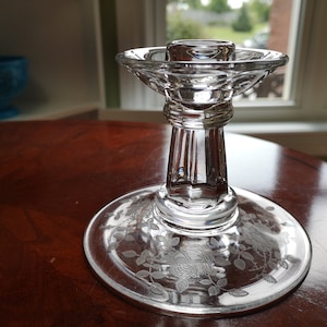 Pode incluir: Um castiçal de vidro transparente com um design gravado decorativo de rosas e folhas. O castiçal tem uma base larga e uma coluna alta e fina.