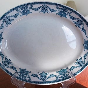 Peut inclure: Un plat de service ovale en céramique blanche avec une bordure décorative bleu foncé. Le rebord est orné d'un motif répétitif composé de fleurs complexes, de vignes entrelacées et de médaillons circulaires avec des dessins centraux.