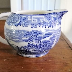 May include: A white ceramic creamer pitcher featuring a blue transferware pattern. The design includes a floral border at the top and a detailed landscape scene with trees, clouds, and buildings around the body. It has a curved handle and a spout.