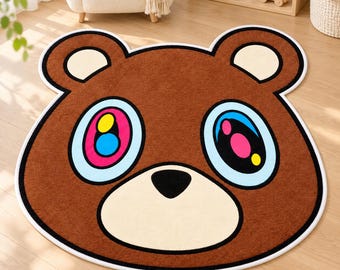 Könnte beinhalten: Ein brauner, bärenförmiger Teppich mit einem großen Kopfdesign, abgerundeten Ohren, einer cremefarbenen Schnauze und einer schwarzen Nase. Die Augen sind mit hellblauen Rändern, pinken Iris und leuchtend gelben und blauen Pupillen gestaltet.