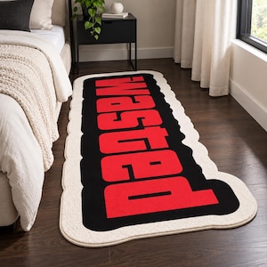 May include: A rectangular area rug with rounded edges featuring a black background and the word WASTED printed in large, bold red capital letters. The rug has a thick, cream-coloured border that follows the scalloped shape of the design.