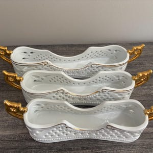 May include: A set of three white porcelain sleigh-shaped dishes featuring a lattice-patterned exterior, gold-painted handles, and gold trim along the top edges. Each elongated vessel has a curved silhouette with openwork detailing on the sides.