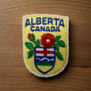 May include: A shield-shaped embroidered patch with a yellow background. The text ALBERTA CANADA is stitched in blue at the top. Below, a red wild rose with green leaves sits above a smaller shield featuring red, white, and blue stripes.