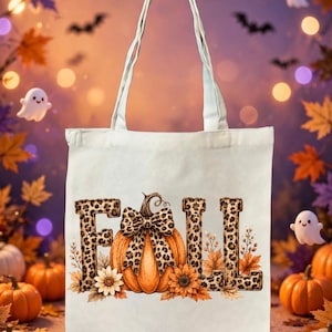 May include: A white canvas tote bag featuring a graphic print of the word FALL in leopard print lettering. The letter A is replaced by an orange pumpkin adorned with a leopard print bow, surrounded by sunflowers and autumn leaves.