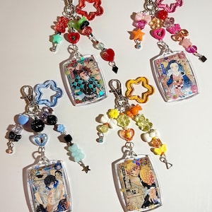May include: Four acrylic keychains featuring anime character illustrations in rectangular frames. Each keychain includes a metal lobster clasp, a colorful star-shaped charm, and various beads in shapes like hearts, bows, and bears in vibrant colors.