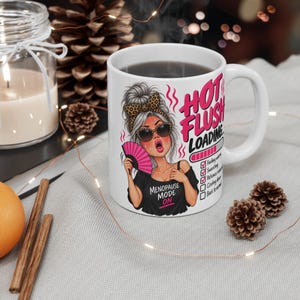 May include: A white ceramic mug featuring a graphic of a person with a messy bun, leopard print headband, and sunglasses, holding a pink hand fan. The text reads HOT FLUSH LOADING with a progress bar and a checklist. The person wears a black shirt with the text MENOPAUSE MODE ON.