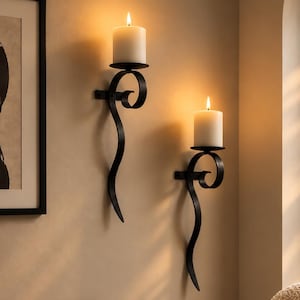 Könnte beinhalten: Zwei schwarze Wandleuchter aus Metall mit geschwungenen, schnörkelartigen Designs, die jeweils eine weiße Stumpenkerze halten. Die Leuchter sind an einer hellen Wand montiert und sorgen für warmes Licht. Die Metallrahmen haben ein dunkles Finish.