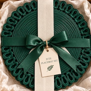 May include: A set of round, dark green jute placemats with a decorative looped edge, presented in a square cardboard gift box lined with white tissue paper. A dark green satin ribbon is tied around the set with a tag labelled JUTE PLACEMAT SET.