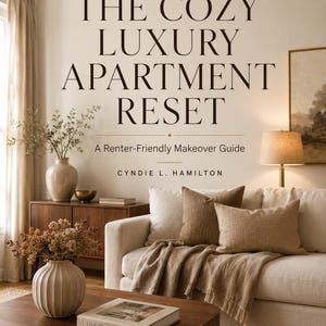 Op de afbeelding: Een boek getiteld THE COZY LUXURY APARTMENT RESET: A Renter-Friendly Makeover Guide door Cyndie L. Hamilton. De cover toont een woonkamer met een beige bank, bruine kussens, een houten salontafel en een boek getiteld LIVE BEAUTIFULLY.