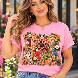 May include: A light pink t-shirt featuring a colourful graphic with the text DOG MAMA. The design includes patchwork patterns, a central paw print, red and white gingham bows, yellow stars, and white daisy flowers with green leaves.