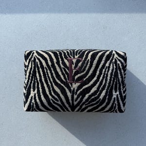 May include: A rectangular zippered pouch featuring a black and white zebra stripe pattern. The center of the pouch is embroidered with a dark maroon capital letter E in a serif font. The fabric has a visible woven texture throughout.