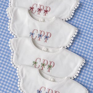 Può includere: Quattro bavaglini bianchi con bordi smerlati e finiture a pompon. Ogni bavaglino presenta le lettere del monogramma CPP ricamate in rosa, accentuate da piccoli fiocchi in filo rosso, blu, verde e blu. I bavaglini hanno una chiusura a pressione.