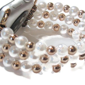 May include: An Apple Watch with a pearl and rose gold beaded band. The band features multiple strands of white faux pearls and smaller rose gold beads. The watch case is silver with a bronze-colored connector.