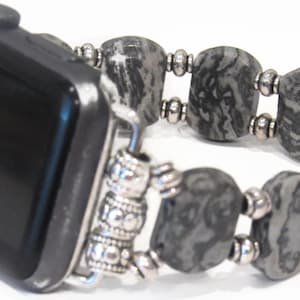 Puede incluir: Reloj inteligente negro con una correa de cuentas grises y plateadas. La correa presenta grandes cuentas de piedra gris estampadas y pequeñas cuentas plateadas. La correa está diseñada para adaptarse a la esfera del reloj.